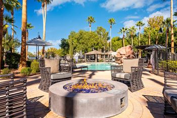A fire pit in the middle of a patio surrounded by chairs and palm trees.